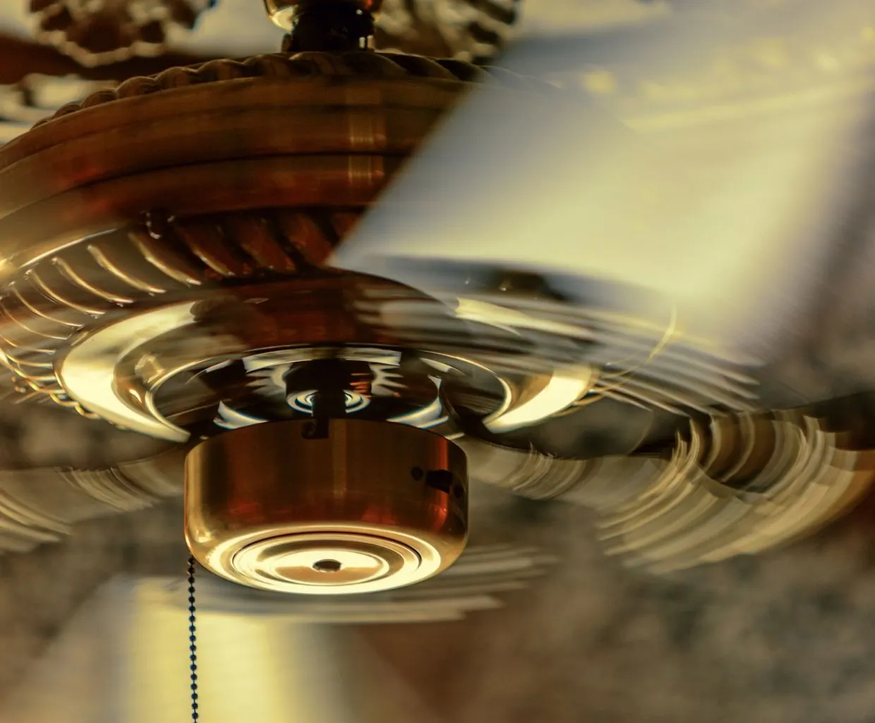 Close-up of a spinning brass ceiling fan motor housing with blurred blades and a pull chain