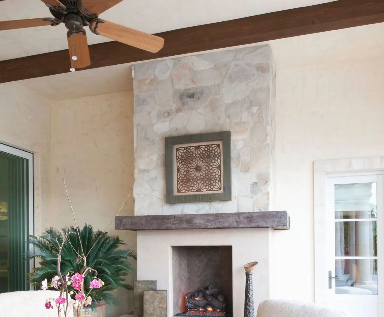 Covered patio with a bronze five-blade ceiling fan with wood blades and pull chains near a stone fireplace