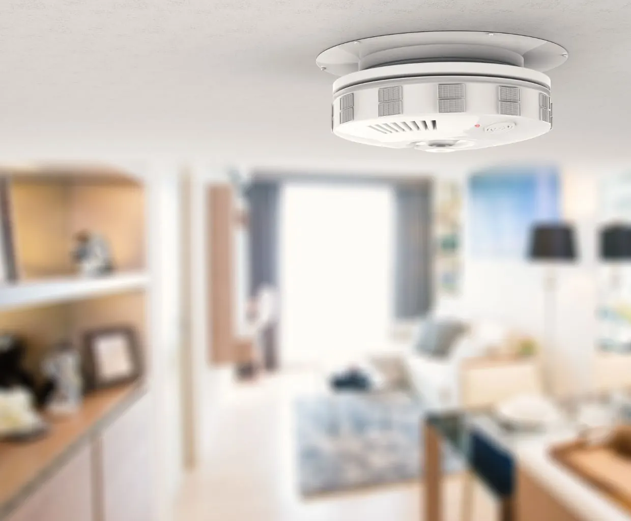 Round white smart device mounted on the ceiling of a bright living room, with blurred shelves and a sofa behind it