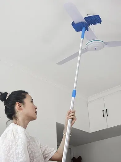 Woman reaching up with a telescoping pole and blue chenille duster head to clean a white ceiling fan blade
