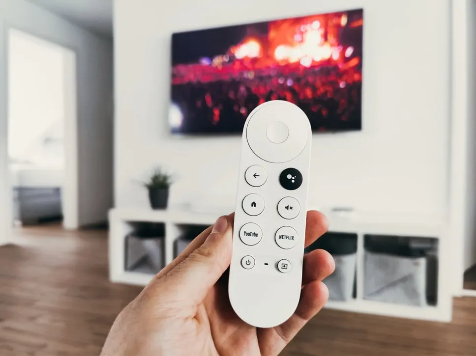 Hand holding a white streaming TV remote in front of a wall-mounted TV in a bright white living room