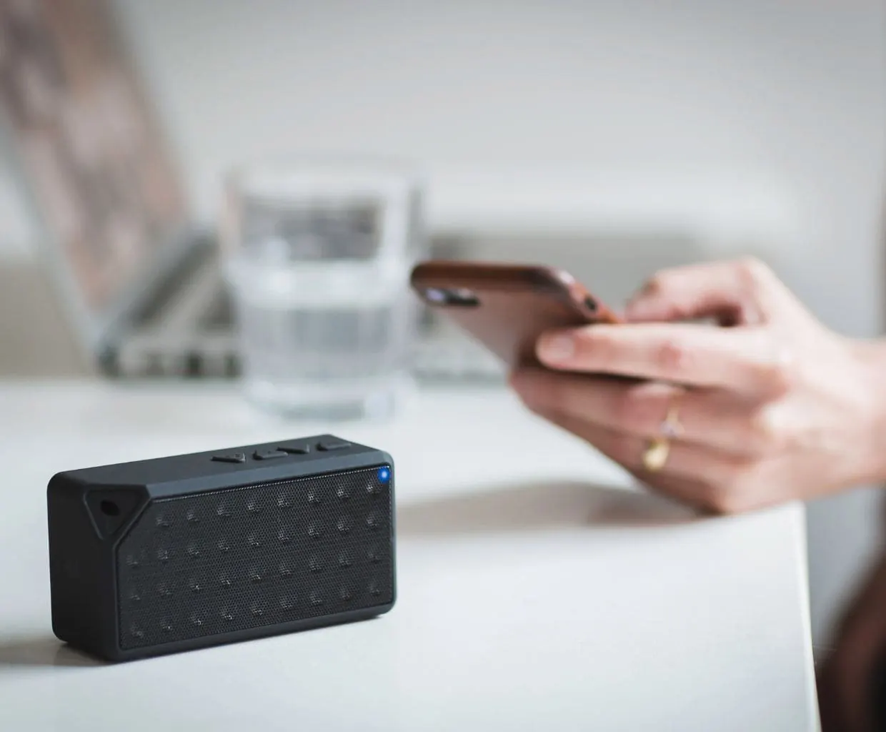 Hand holding a smartphone beside a small black wireless speaker with a blue light on a white table