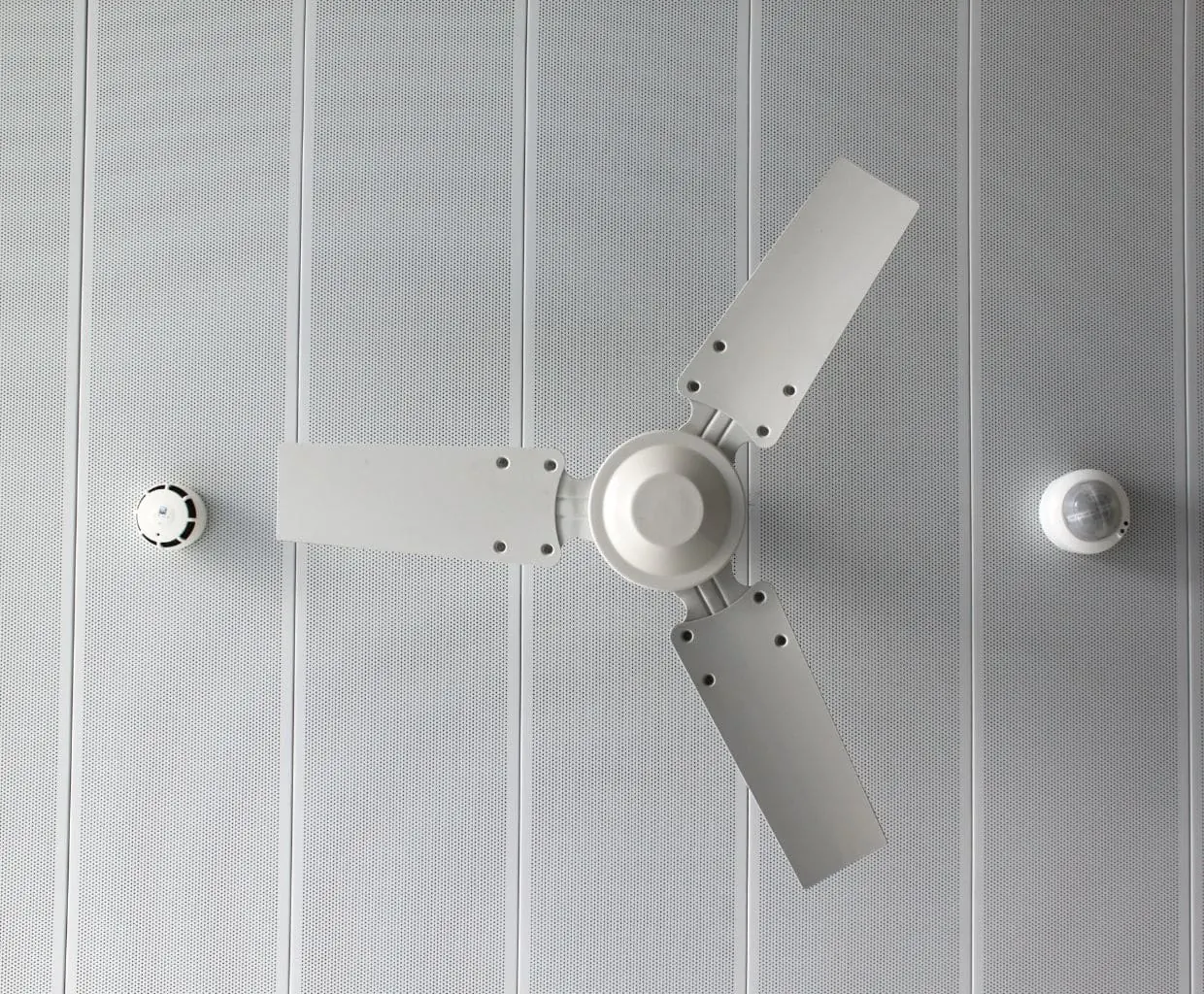 White three-blade ceiling fan with no light kit on a perforated white panel ceiling between two round ceiling sensors