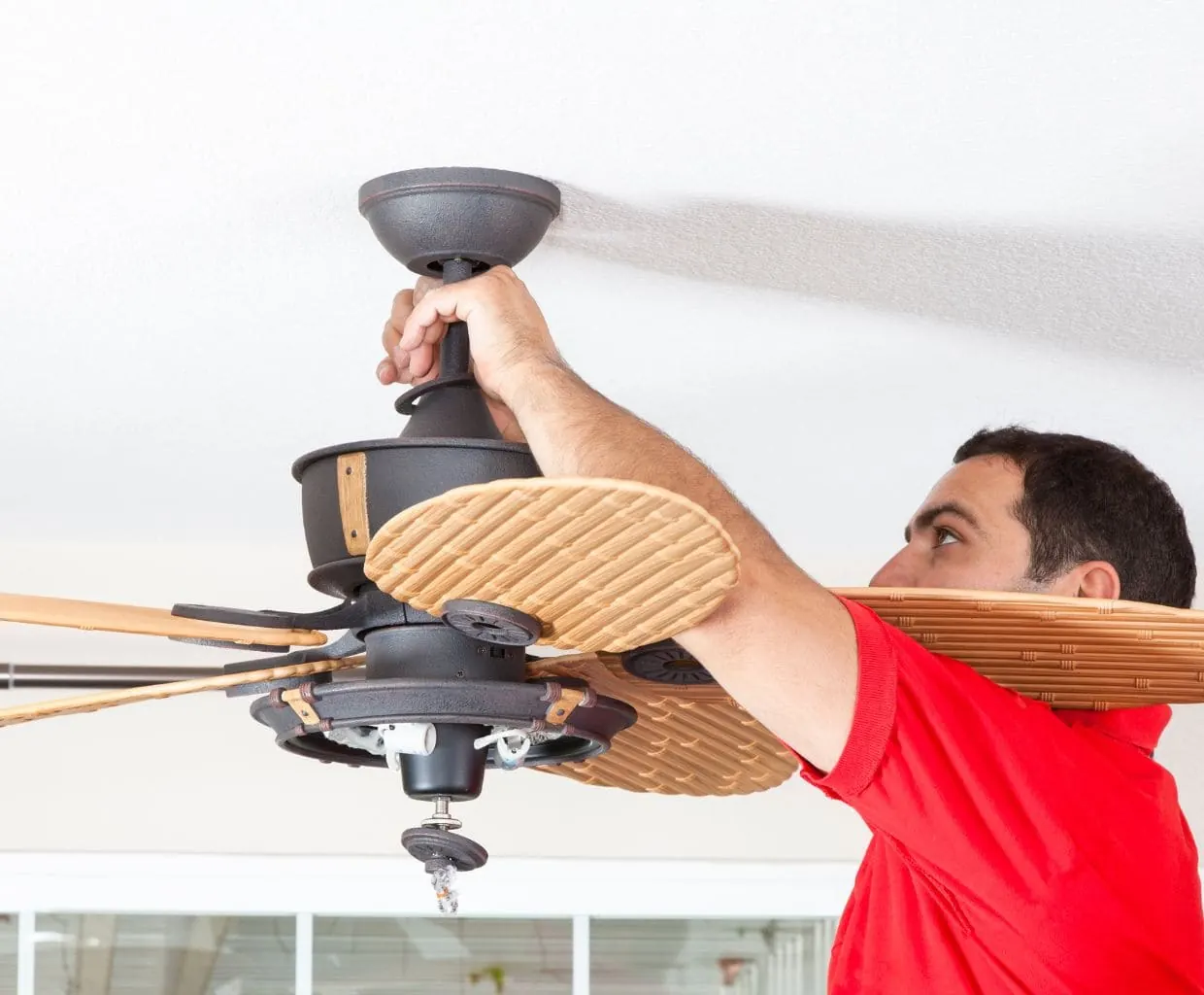 Man in a red shirt holding the downrod of a dark bronze ceiling fan with woven wicker-look blades