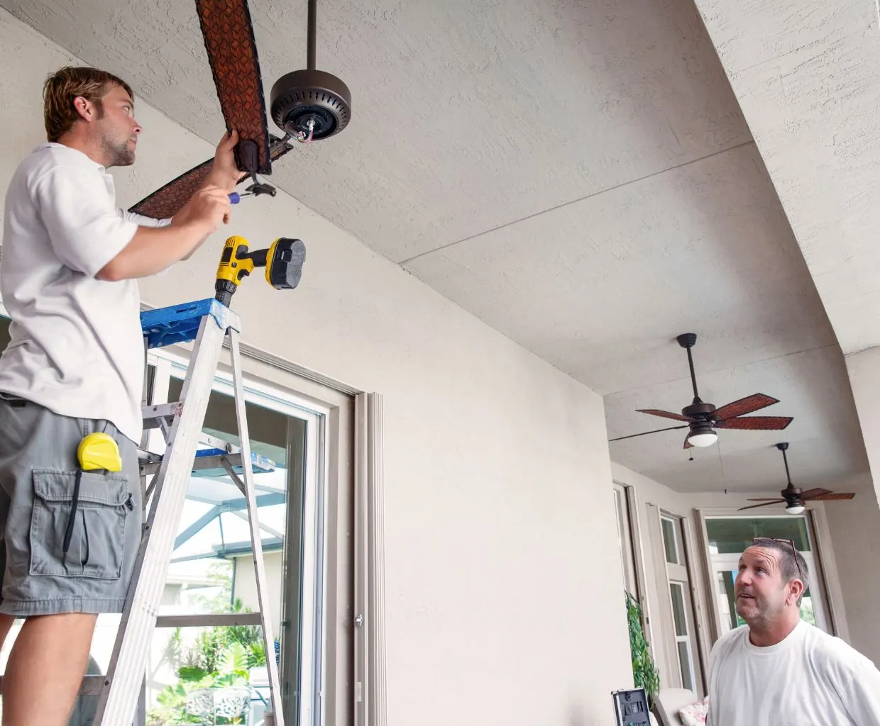 Man on a ladder installing a dark ceiling fan on a covered patio as a coworker looks up, two fans already hung behind