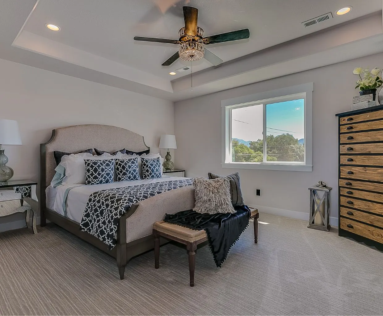 Bedroom with a tray ceiling and a five-blade ceiling fan with a crystal light above an upholstered bed and bench
