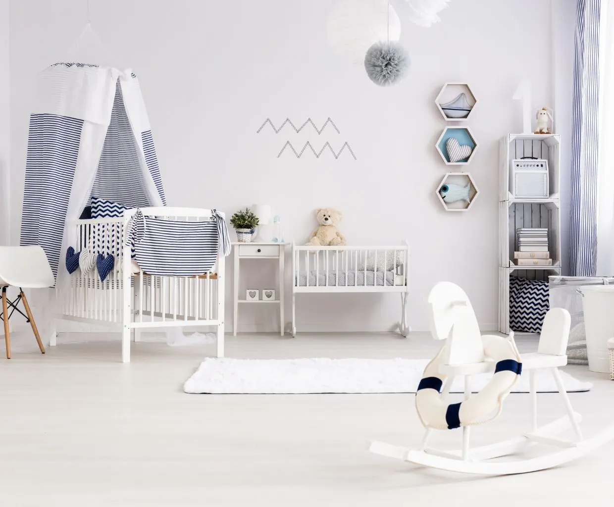 Bright white nursery with a canopy crib, a small cradle with a teddy bear, hexagon wall shelves and a rocking horse