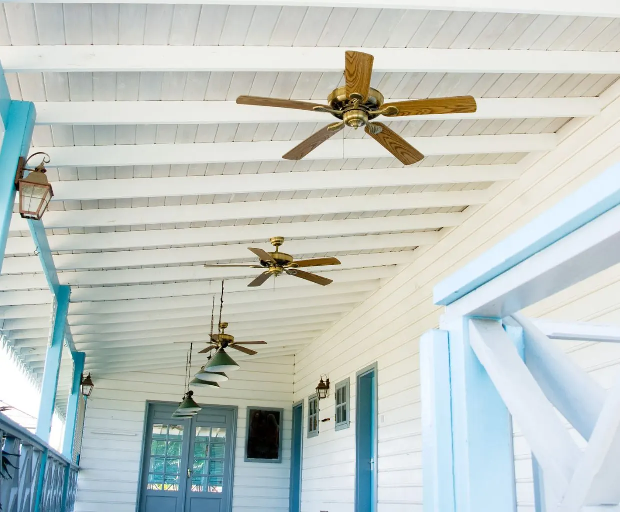 Covered white porch with three brass ceiling fans with wood blades, light-blue posts and hanging pendant lamps