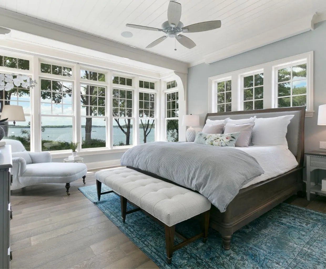 Bright bedroom with a white five-blade ceiling fan over an upholstered bed, tufted bench and windows facing a lake